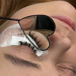Heart-shaped lash mirror reflecting a client's eyelash extensions during a lash application, with under-eye pads in place.