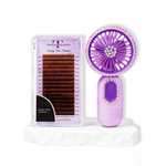 Purple portable lash fan with rhinestone detailing displayed beside a lash extension tray on a white stone surface.