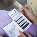 Lash artist holding a marble-pattern tweezer above a lash extension tray with multiple lash styles arranged in a case.