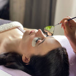 Lash artist using a heart-shaped mirror to check a client’s eyelash extensions during a lash appointment, with an under-eye pad in place.