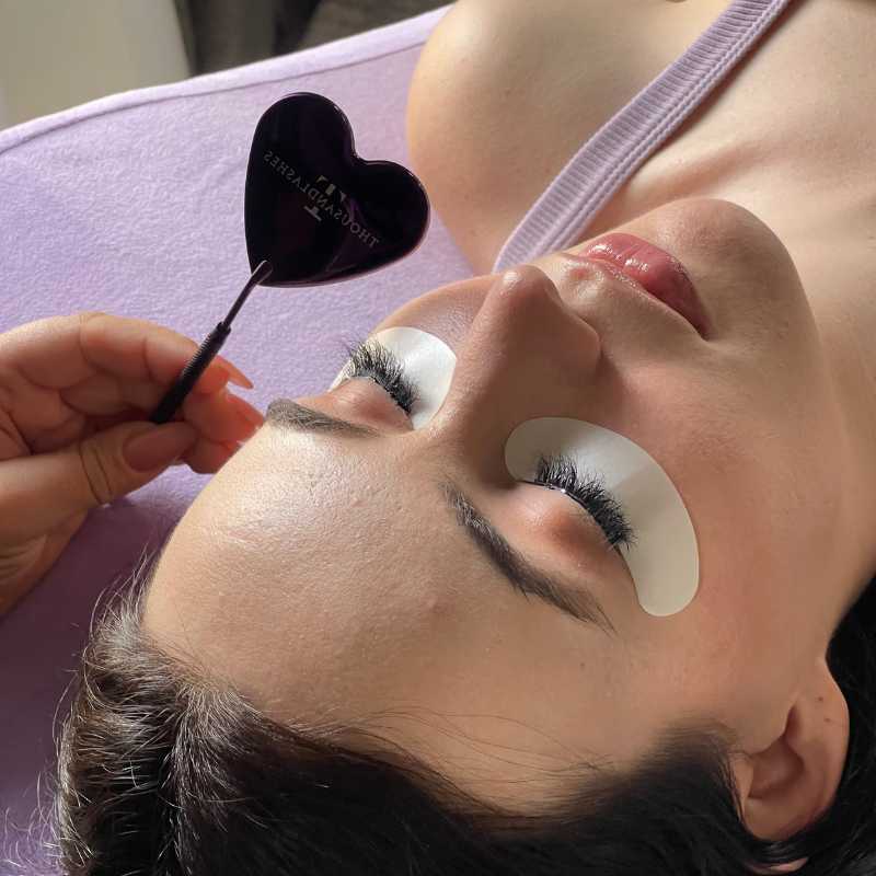 Lash artist using a purple heart-shaped mirror to check a client’s eyelash extensions, with under-eye gel pads in place.